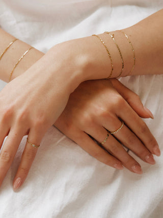 The Bond Society gold stacking rings and delicate chain bracelets on woman's hands, minimalist fine jewelry styled on white fabric.