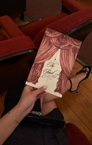 The Bond Society delicate gold bracelet styled on woman’s wrist while holding a theater program inside a red velvet seat venue.