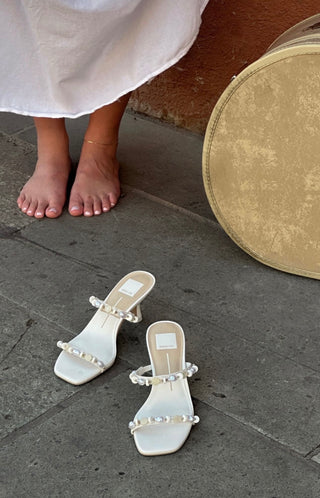The Bond Society minimalist gold anklet styled on bare ankle with white dress, paired with pearl-embellished sandals on stone pavement
